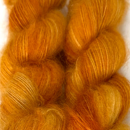 Delicious by Apple Fiber Studio. Close-up of two twisted skeins of fluffy yarn in a warm golden orange color with subtle lighter yellow highlights, set against a plain light background.