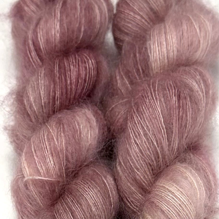 Delicious by Apple Fiber Studio. Two twisted skeins of fluffy mohair yarn in a dusty mauve-purple color with subtle silver-white highlights, set against a plain white background.