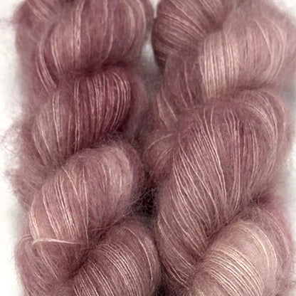Delicious by Apple Fiber Studio. Two twisted skeins of fluffy mohair yarn in a dusty mauve-purple color with subtle silver-white highlights, set against a plain white background.