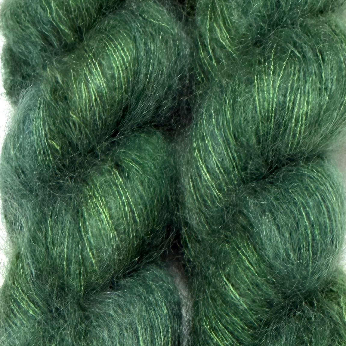 Delicious by Apple Fiber Studio. Close-up of two fluffy skeins of fuzzy yarn in a deep forest green color with fine lime green and yellow shimmering threads.