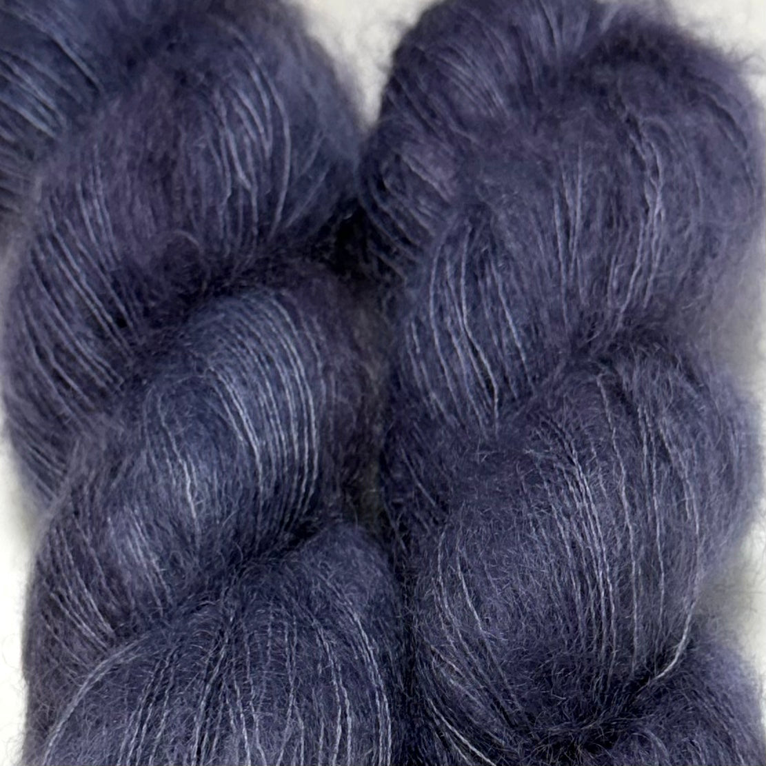 Delicious by Apple Fiber Studio. Close-up of two soft, fuzzy yarn skeins in a dark muted blue-violet tone with subtle lighter streaks, set against a light neutral background.