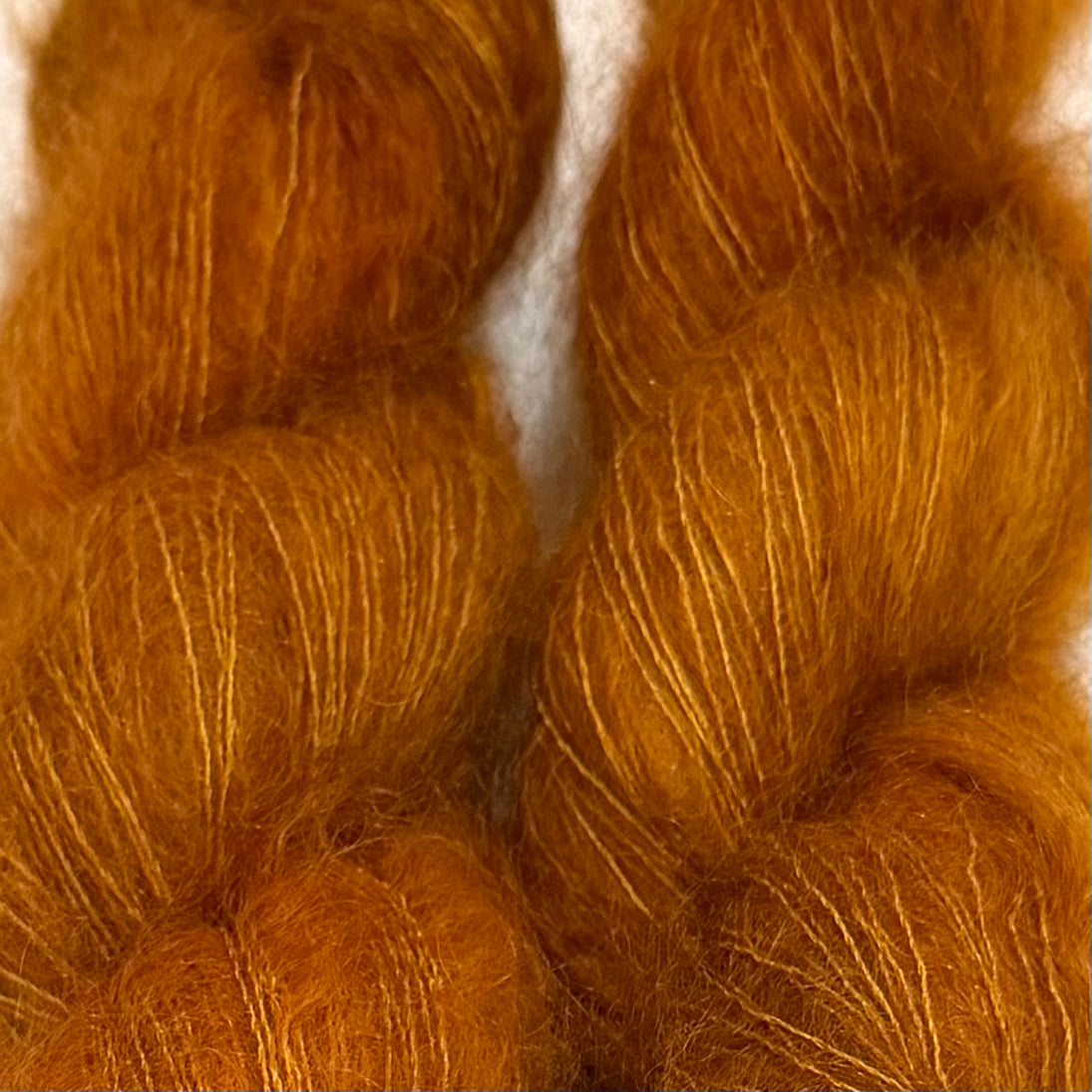Delicious by Apple Fiber Studio. Close-up of two skeins of fluffy yarn in a warm golden orange-brown color, filling the frame against a small white background.