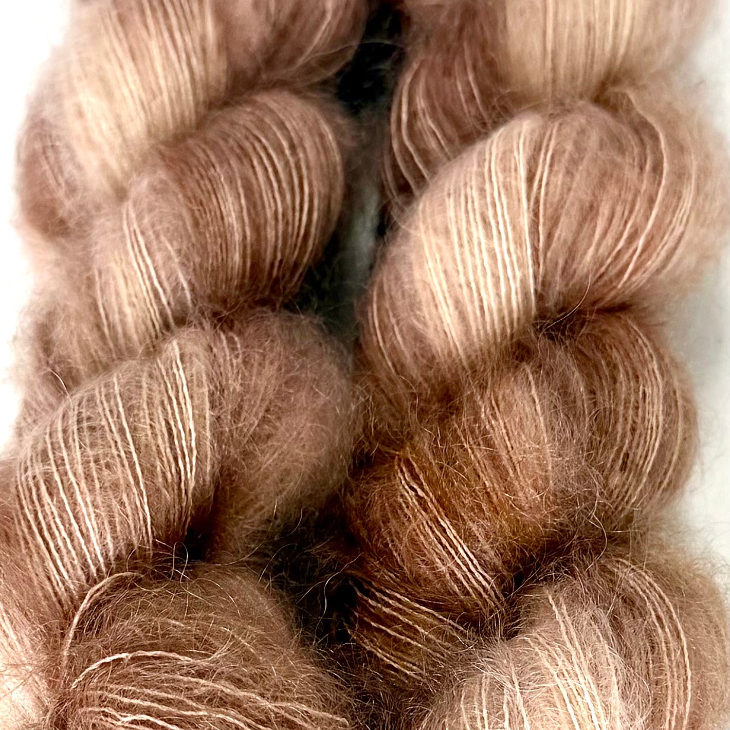 Delicious by Apple Fiber Studio. Close-up of two twisted skeins of fluffy yarn in a warm taupe-brown color with lighter cream highlights, shown against a plain light background.