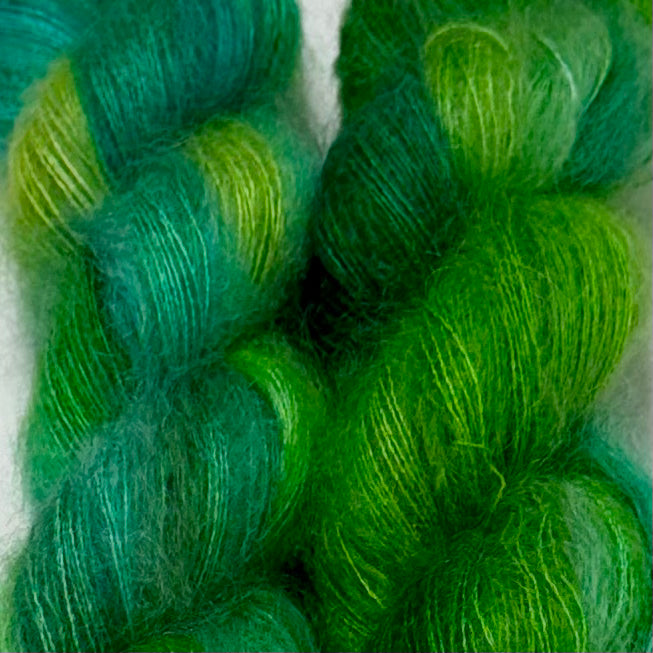 Delicious by Apple Fiber Studio. Close-up of two skeins of fuzzy yarn in shades of green and teal, showing the soft texture and variegated color from bright lime green to deep emerald and teal.