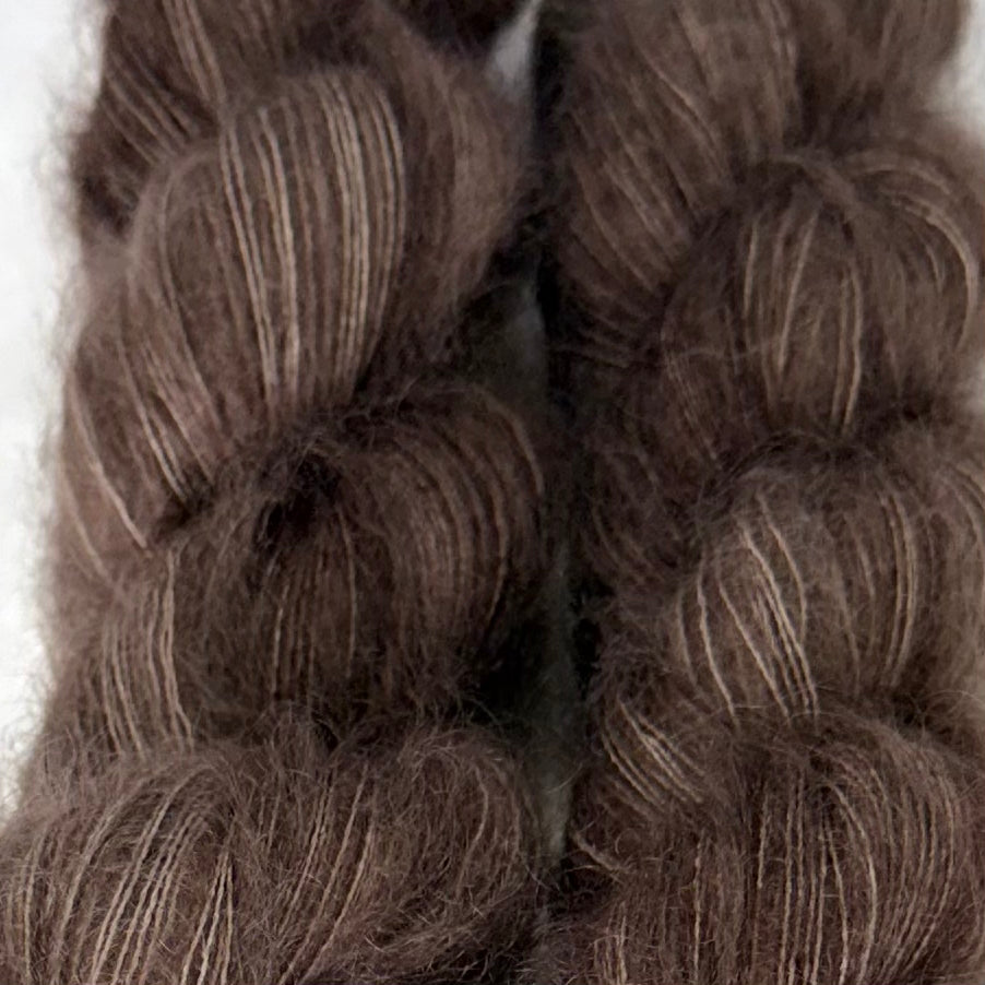 Delicious by Apple Fiber Studio. Close-up of two braided yarn skeins in a dark brown color with subtle light beige highlights, showing the soft texture of the fibers.