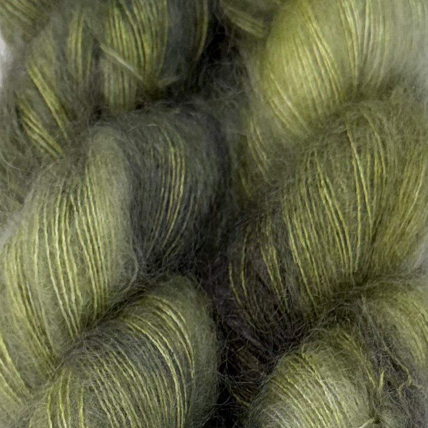 Delicious by Apple Fiber Studio. Close-up of fuzzy yarn skeins in a muted olive green and dark gray colorway, showing the soft texture and subtle sheen of the fibers.