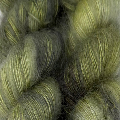 Delicious by Apple Fiber Studio. Close-up of fuzzy yarn skeins in a muted olive green and dark gray colorway, showing the soft texture and subtle sheen of the fibers.