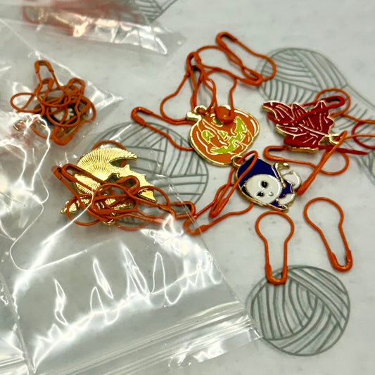 Holiday Stitch Markers - Several small enamel lapel pins attached to orange safety pins, arranged on a light surface with yarn balls and a clear plastic bag in the background.