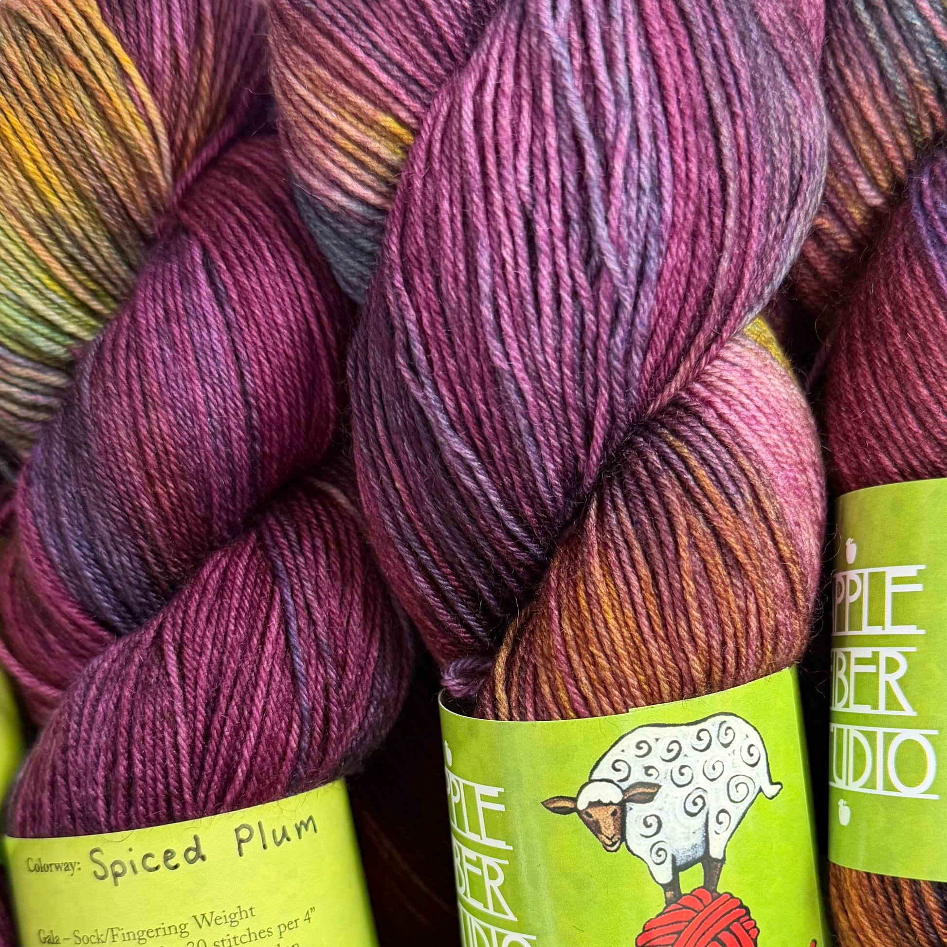 YarnFall 2026 Spiced Plum Hand-Dyed Yarn - Sock & Worsted, Close-up of several skeins of yarn in a variegated plum, purple, pink, orange, and yellow blend. A bright green paper label wraps around the front skein, with the brand name 'Pifaber Studio' and a sheep illustration visible.
