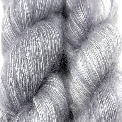 Delicious by Apple Fiber Studio. Close-up of a twisted skein of fluffy yarn in a soft heathered gray and white blend, showing the fuzzy fiber texture against a light neutral background.