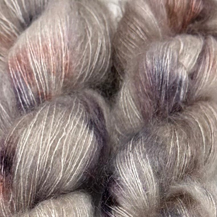 Delicious by Apple Fiber Studio. Close-up of soft, fuzzy yarn skeins in a heathered blend of gray, beige, silver, and muted mauve-purple tones.