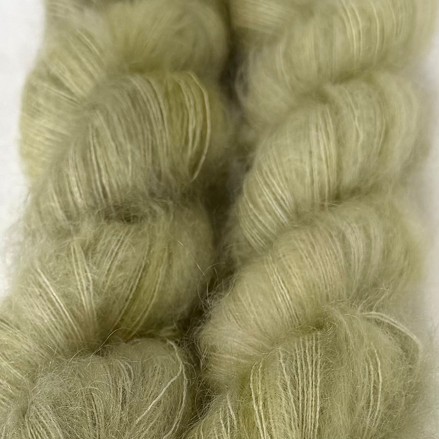 Delicious by Apple Fiber Studio. Close-up of two soft, fuzzy skeins of pale sage green yarn against a light neutral background.