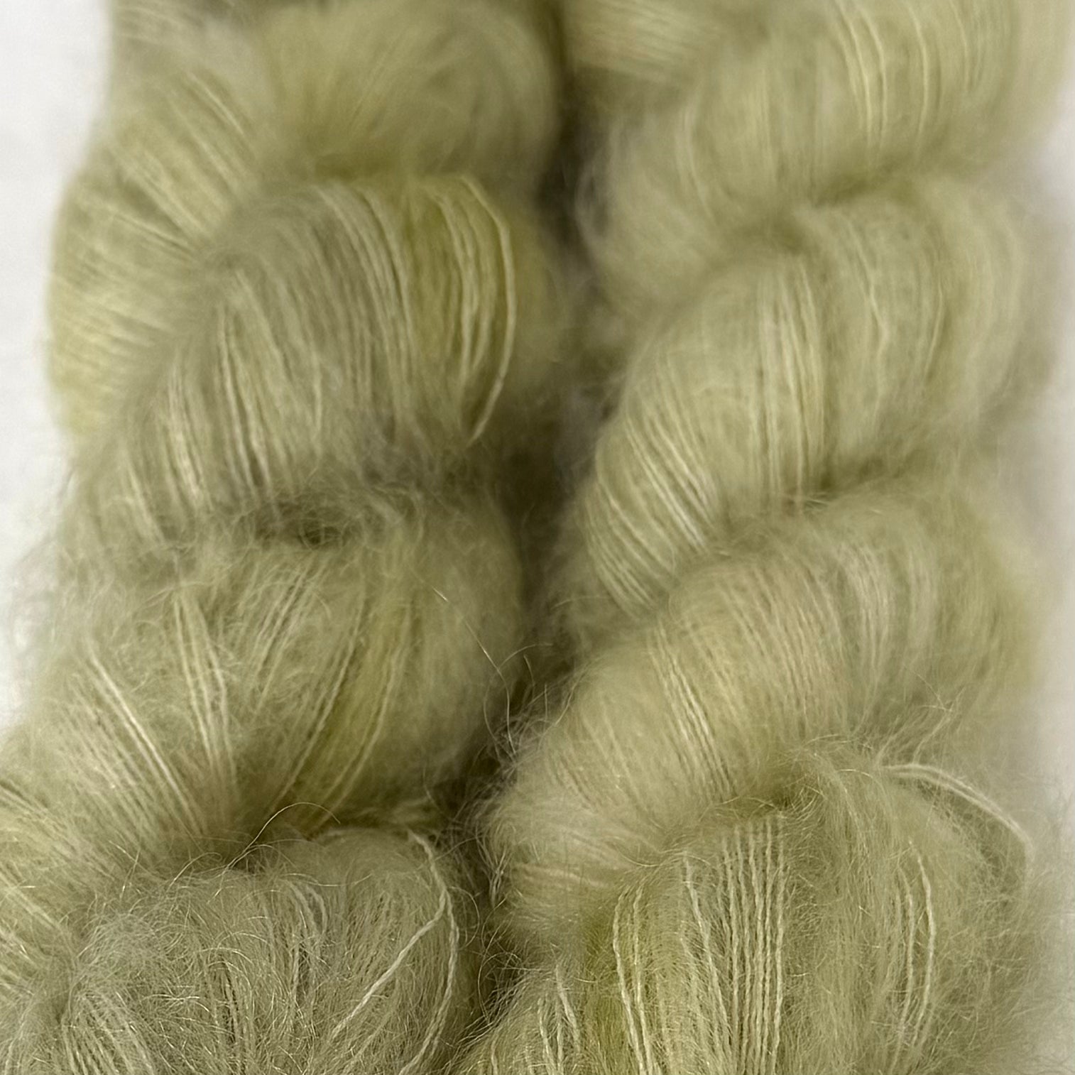 Delicious by Apple Fiber Studio. Close-up of two soft, fuzzy skeins of pale sage green yarn against a light neutral background.