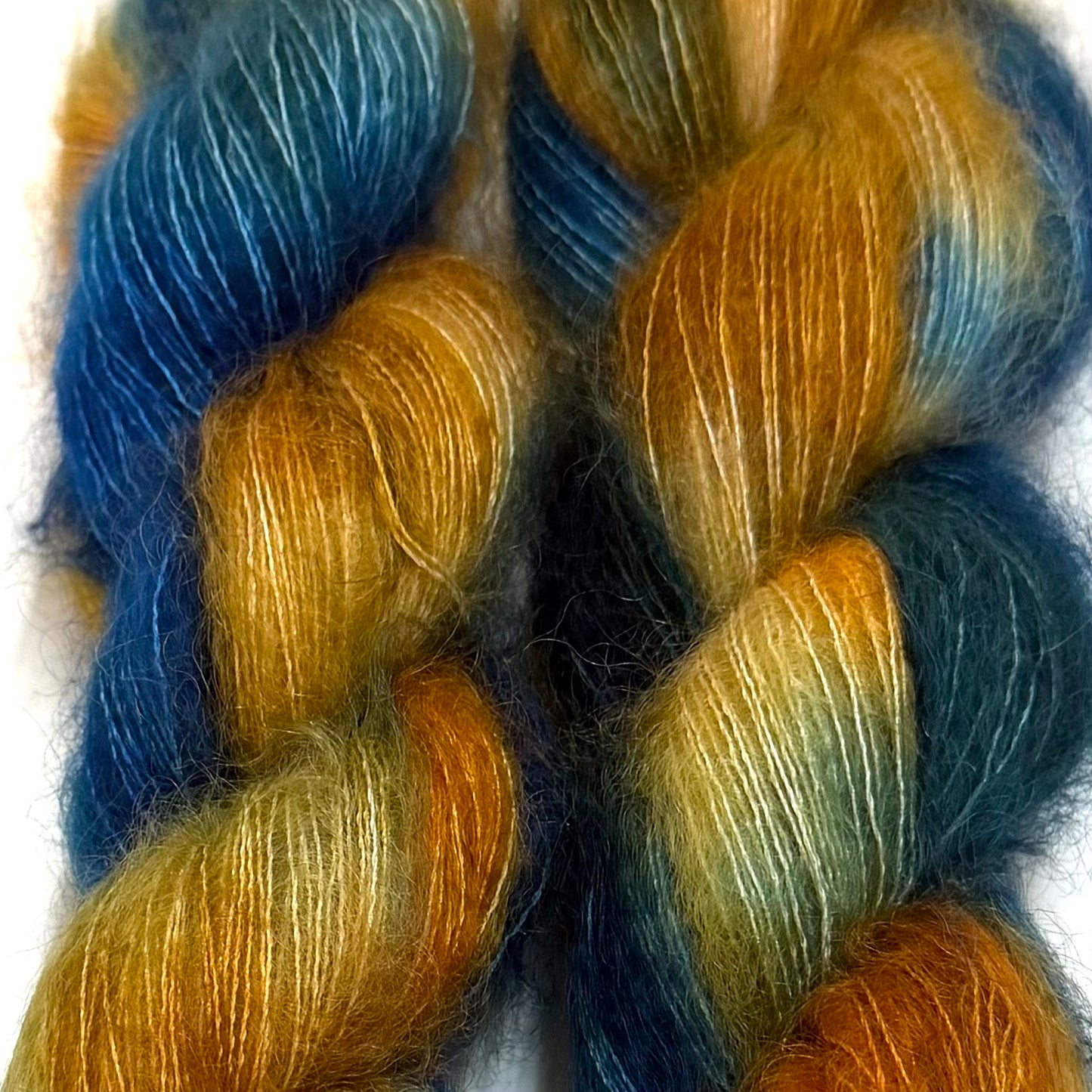 Delicious by Apple Fiber Studio. Close-up of two twisted skeins of fluffy yarn with variegated colors of deep blue, mustard gold, teal, and orange on a white background.
