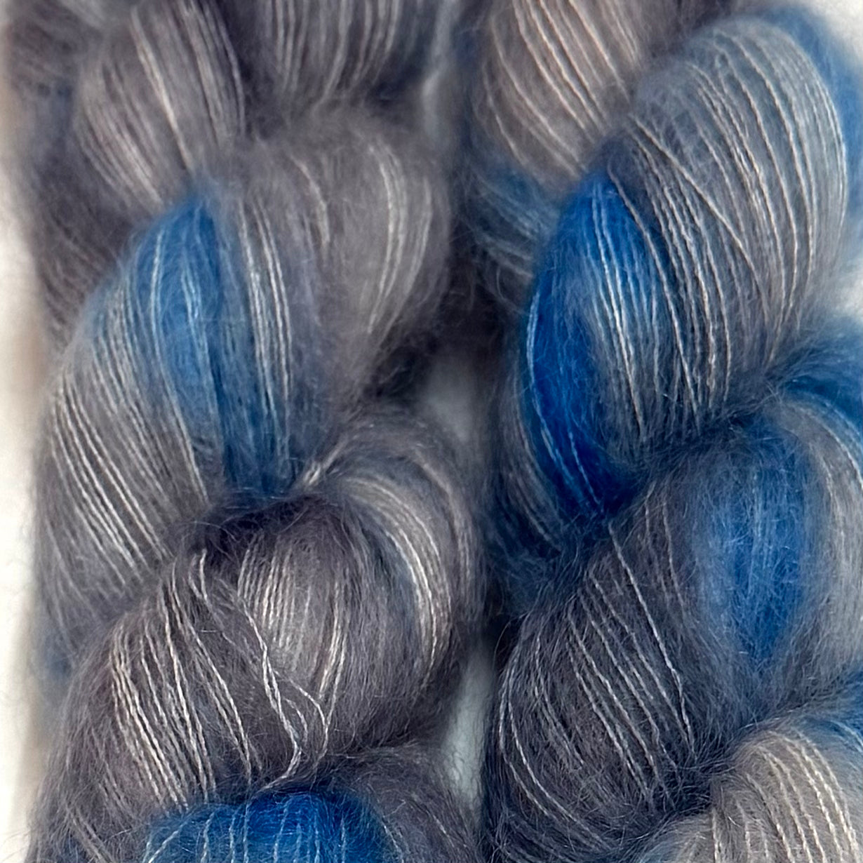 Delicious by Apple Fiber Studio. Close-up of two twisted skeins of fuzzy yarn in a heathered gray and silver tone with vivid blue sections, photographed against a light neutral background.