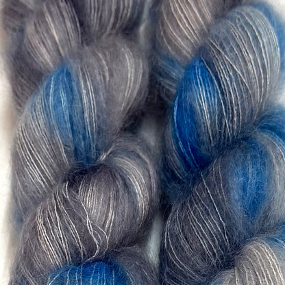 Delicious by Apple Fiber Studio. Close-up of two twisted skeins of fuzzy yarn in a heathered gray and silver tone with vivid blue sections, photographed against a light neutral background.