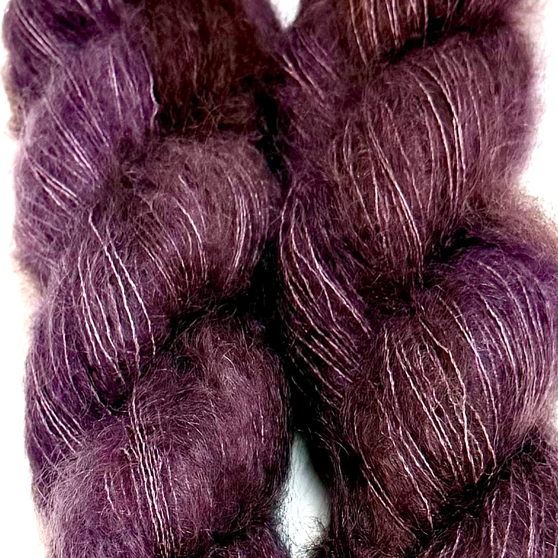 Delicious by Apple Fiber Studio. Two twisted skeins of fuzzy yarn in a deep plum-purple color with fine silver-white highlights, shown against a plain white background.